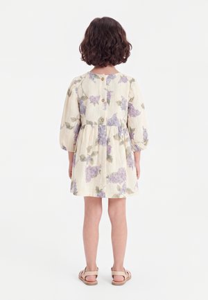 Enfant aux cheveux courts et bouclés portant une robe crème à imprimé floral violet avec des boutons dans le dos, debout de dos en sandales beiges.