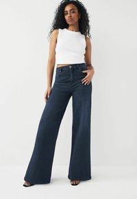 Bredde ben mørke denim jeans med høj talje, der har et klassisk fem-lomme-design og en diskret syningdetalje. Kombineret med en hvid ærmeløs top.