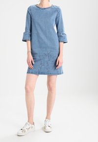 Denimmekko, jossa on kolmanneksen mittaiset levenevät hihat, pyöreä pääntie ja kukkakirjonta helman kohdalla. Yhdistetty valkoisiin tennareihin, joissa on kuvioidut yksityiskohdat.