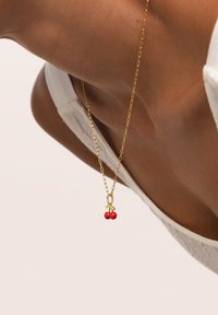 Collar de cadena de oro con un pequeño colgante de cereza llevado en el cuello de una persona que viste una prenda sin mangas de textura blanca.
