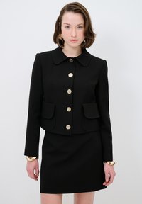 Zwarte op maat gemaakte blazer met grote kraag, gouden knopen en opgestikte zakken. Gecombineerd met een bijpassende fitted rok, met een gladde textuur.