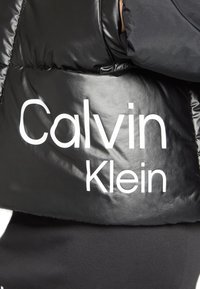Svart puffjacka med glänsande yta, med vit "Calvin Klein" logotyp på sidan och en figursydd design.