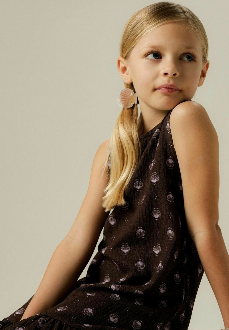 Robe marron foncé avec des accents en forme de coquillage, fabriquée en tissu texturé. Le modèle a une queue de cheval blonde claire et porte une boucle d'oreille en forme de coquillage.