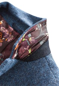 Collo in tweed di lana blu con motivo a spina di pesce; foderato in floreale con tonalità bordeaux scuro e accenti verdi. Texture morbida e finitura su misura.