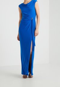 Long, blue dress with a draped neckline, side slit, and gathered detail at the waist featuring a decorative accent for added flair.