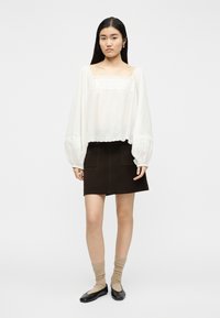 Jeune femme portant un chemisier blanc à manches longues avec des détails en dentelle, une jupe marron foncé avec des poches avant, des chaussettes beiges et des ballerines noires, debout.