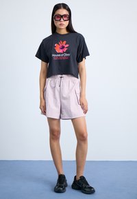 Tricou scurt negru cu grafică florală roșie, pantaloni scurți roz deschis cu talie elastică și buzunare laterale, și pantofi sport negri.