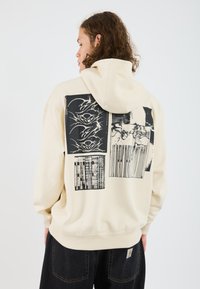 Felpa crema con grafiche nere stampate sul retro, caratterizzate da design astratti. Tessuto morbido, vestibilità comoda e cappuccio regolabile.