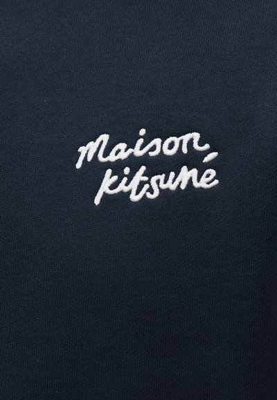Λευκό κεντημένο κείμενο "Maison Kitsuné" με γραφή σε καλλιγραφικό στιλ σε σκούρο μπλε ύφασμα.
