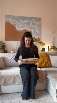 Wełniany pled narzucony na jasno szarą kanapę. Kobieta w czarnym swetrze i szarych spodniach trzyma ozdobną poduszkę z napisem.