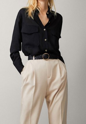 Femme portant une chemise noire à boutons rentrée dans un pantalon beige taille haute plissé avec une ceinture noire tressée.