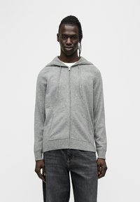 Hoodie gris à fermeture éclair fabriqué en tissu doux, avec une capuche à cordon, des poches avant et un ourlet côtelé. Porté avec un jean denim foncé.