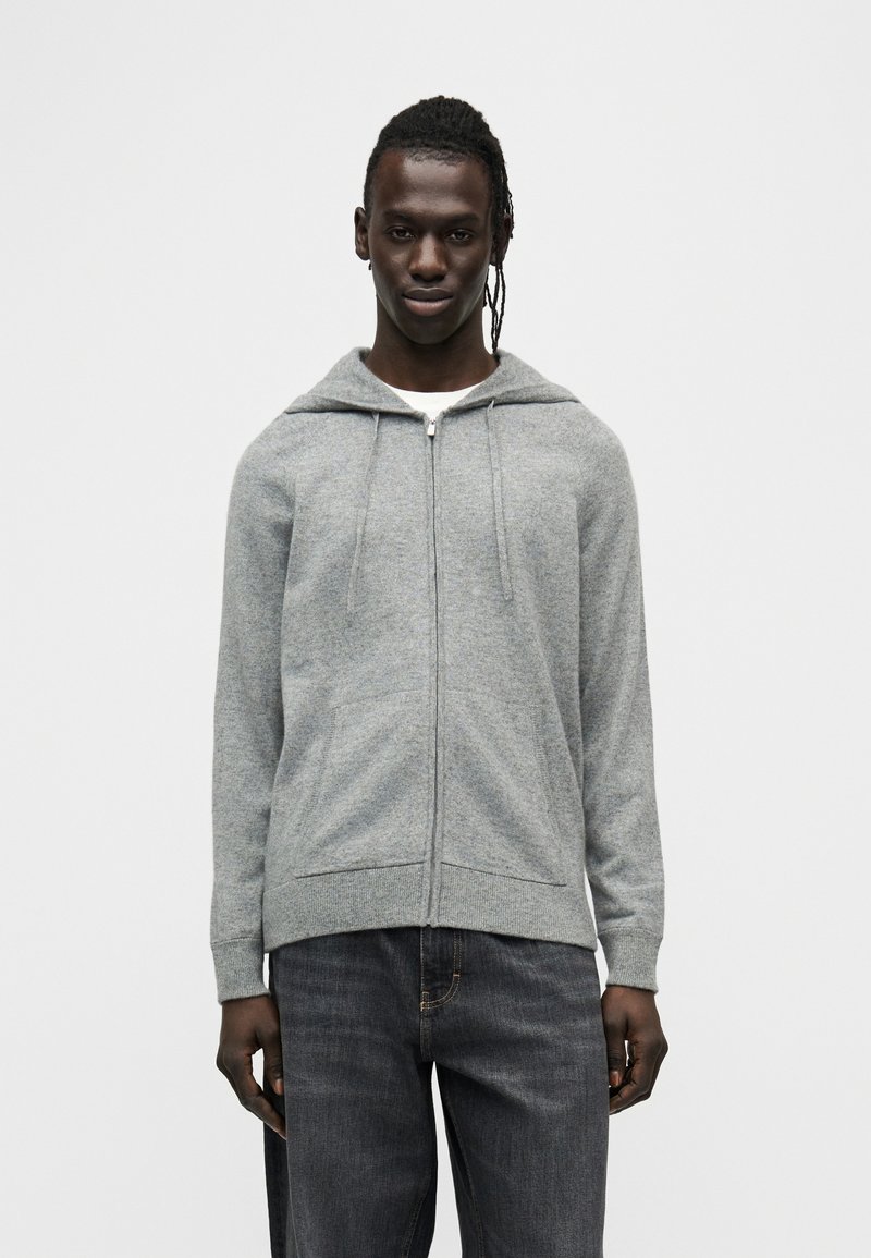 Hoodie gris à fermeture éclair fabriqué en tissu doux, avec une capuche à cordon, des poches avant et un ourlet côtelé. Porté avec un jean denim foncé.