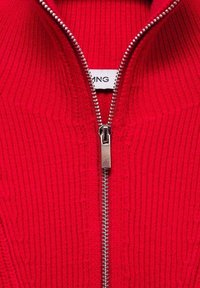 Primo piano di un indumento a maglia rossa a coste con una cerniera metallica argento parzialmente chiusa e un'etichetta di abbigliamento ben visibile all'interno.