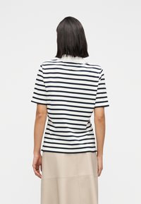 Γυναίκα με ίσια σκούρα μαλλιά φοράει μια κοντομάνικη μπλούζα με άσπρες και μαύρες ρίγες και μια μπεζ φούστα, γυρισμένη πλάτη, σε απλό φόντο.