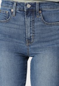 Traperice visokog struka u plavoj boji s izblijedjelim pranjem, s prednjim džepovima, metalnim gumbom i kontrastnim šavovima.