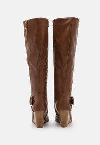 Paire de bottes montantes en cuir marron avec talons compensés, fermetures éclair latérales et sangles à boucle, vues de dos sur fond blanc.