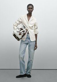 Creamfarbener Blazer mit einem einzigen Knopf, hellblaue Jeans und eine braun-weiß gemusterte Handtasche. Das Model steht vor einem neutralen Hintergrund.