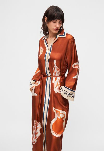 Γυναίκα με φόρεμα σε σκουριά απόχρωση, με πορτοκαλί και λευκά διακοσμητικά μοτίβα με φύλλα, κουμπιά μπροστά και ζώνη στη μέση, κοιτάζει προς τα κάτω.