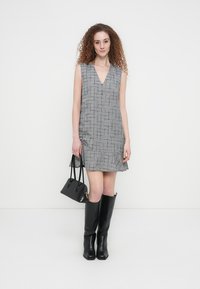 Mujer con cabello rizado y rojo, vestida con un vestido sin mangas de cuadros grises, botas negras hasta la rodilla y sosteniendo un bolso negro sobre un fondo blanco.