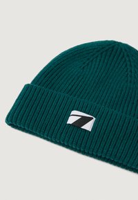 Gorro de punto en color verde azulado con una textura de canalé ancha y un puño doblado. Presenta un parche blanco con un logo de franja diagonal negra.