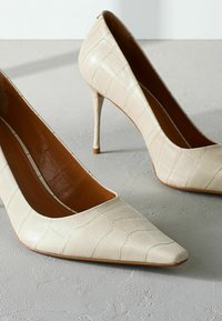 Décolleté in finta pelle beige con punta appuntita e tacco a stiletto, caratterizzate da una texture simile a quella di un coccodrillo e un interno liscio.