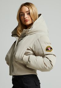 Beige pufferjack met hoge kraag, geribbelde manchetten en een badge op de linkermouw. Voorzien van een ritssluiting aan de voorkant en een zachte textuur.