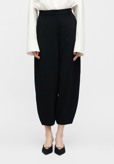 Γυναίκα που φοράει μαύρο παντελόνι υψηλής μέσης με φαρδύ cropped πόδι, λευκό πουκάμισο με μακριά μανίκια και μαύρα παπούτσια μυτερά.