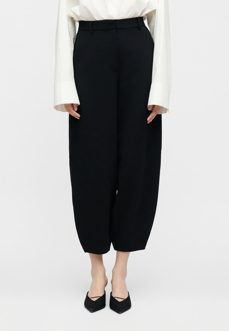 Frau trägt schwarze, hoch taillierte, weite, knöchellange Hosen mit einer weißen Langarmbluse und schwarzen spitz zulaufenden Schuhen.