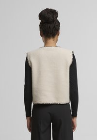 Béžová fleecová vesta s texturovaným povrchem a leopardím vzorem. Bez rukávů, kulatý výstřih, pohled ze zad.