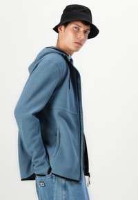 Kék fleece dzseki textúrázott mintázattal az oldalán, elöl cipzárral és kéz zsebekkel; fekete vödörkalappal és világos farmernadrággal párosítva.