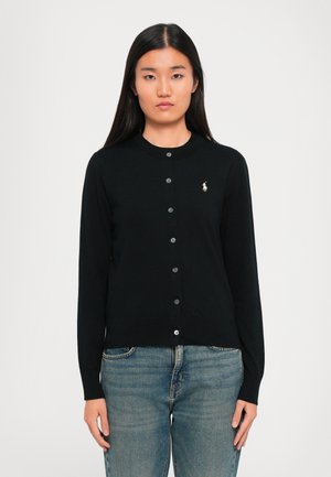 Frau mit langen schwarzen Haaren, die einen schwarzen Knopf cardigan mit einem kleinen weißen Logo auf der Brust und blauen Jeans trägt, frontal ausgerichtet.