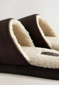 Brauner Hausschuh mit einer weichen, cremefarbenen Innenauskleidung. Er bietet ein glattes, abgerundetes Design und eine strukturierte Außenseite für besseren Halt.