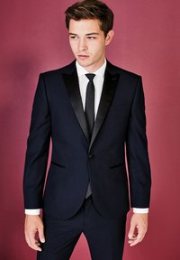 Completo blu navy con revers in satin e taglio slim, abbinato a una camicia bianca e una cravatta nera. Caratterizzato da tasche laterali e una silhouette sapientemente sagomata.