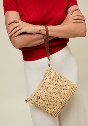 Henkilö, joka pitää punaista yläosaa ja valkoisia housuja, pitää beigeä kudottua laukkua, jossa on ruskea nahkahihna, kädet ristissä rinnan päällä.