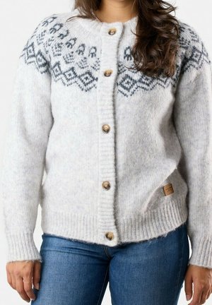 Femme portant un cardigan en maille gris clair avec des motifs géométriques noirs sur la poitrine et des boutons en bois, associé à un jean bleu.