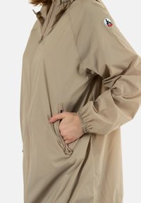 Chaqueta ligera beige con cierre frontal, puños elásticos y bolsillos laterales. Presenta un parche con el logo en la parte superior del brazo. Textura suave.