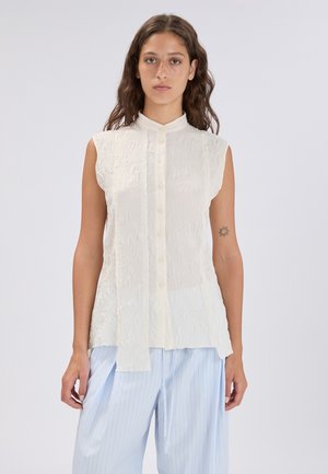 Giovane donna con capelli castani ondulati, che indossa una camicetta bianca senza maniche con bottoni e una texture, abbinata a pantaloni larghi a righe sottili azzurri.