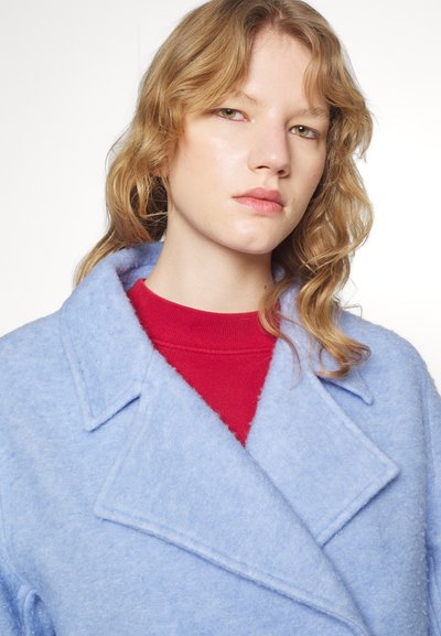 Manteau en mélange de laine bleu clair avec un large col, superposé à une chemise rouge. Le manteau présente une texture douce et une coupe ample, soulignant l'élégance décontractée.