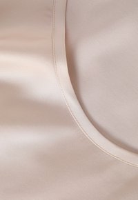 Beżowa satynowa tkanina o gładkiej fakturze, z zaokrąglonym dekoltem i czystym, szytym brzegiem. Subtelny blask widoczny na powierzchni.