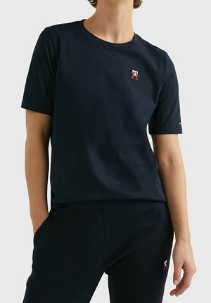 Personne portant un T-shirt bleu marine à manches courtes et un pantalon assorti, tous deux avec un petit logo brodé rouge et blanc sur le côté gauche.