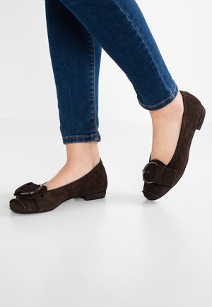 Fødder iført mørkebrune ruskind sko med lave hæle og store runde spænder, kombineret med skinny blå jeans mod en hvid baggrund.