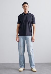 Marineblå polo t-skjorte med hvit kragedetalj, kombinert med lyseblå avslappede jeans. Modellen står på grått gulv mot en ensfarget bakgrunn.