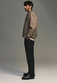 Veste bomber marron texturée avec des poignets côtelés, portée sur une chemise blanche et un pantalon sombre ; associée à des chaussures noires à semelle épaisse.