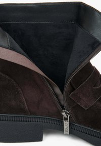 Detailní záběr na tmavě hnědou semišovou botu s černou podešví, uvnitř s měkkou černou podšívkou a částečně otevřeným zipem, který odhaluje kovový táhlo s vyrytým názvem značky.