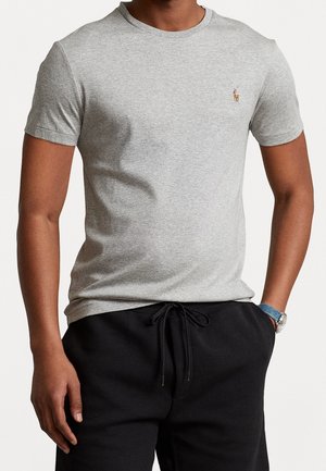 Uomo che indossa una t-shirt a maniche corte grigio chiaro con scollo rotondo e un piccolo logo ricamato sul petto, abbinata a pantaloni neri con coulisse.
