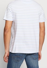 Persona vistiendo una camisa de manga corta blanca y azul claro con rayas horizontales y jeans de mezclilla azules, vista desde la espalda.