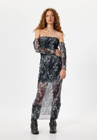 Abito nero senza spalline con maniche lunghe trasparenti, con motivi floreali e di testo, design arricciato e un lungo orlo trasparente; abbinato a stivaletti neri alla caviglia.
