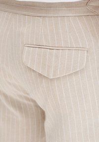 Beżowe materiałowe spodnie w paski z jedną tylną kieszenią z ostro zakończonym klapkiem. Tekstura wydaje się gładka i lekka.