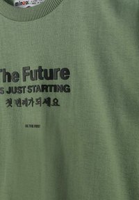 Vêtement vert avec texte noir disant « L'avenir ne fait que commencer » et des caractères coréens, ainsi qu'un texte plus petit « Soyez le premier ».